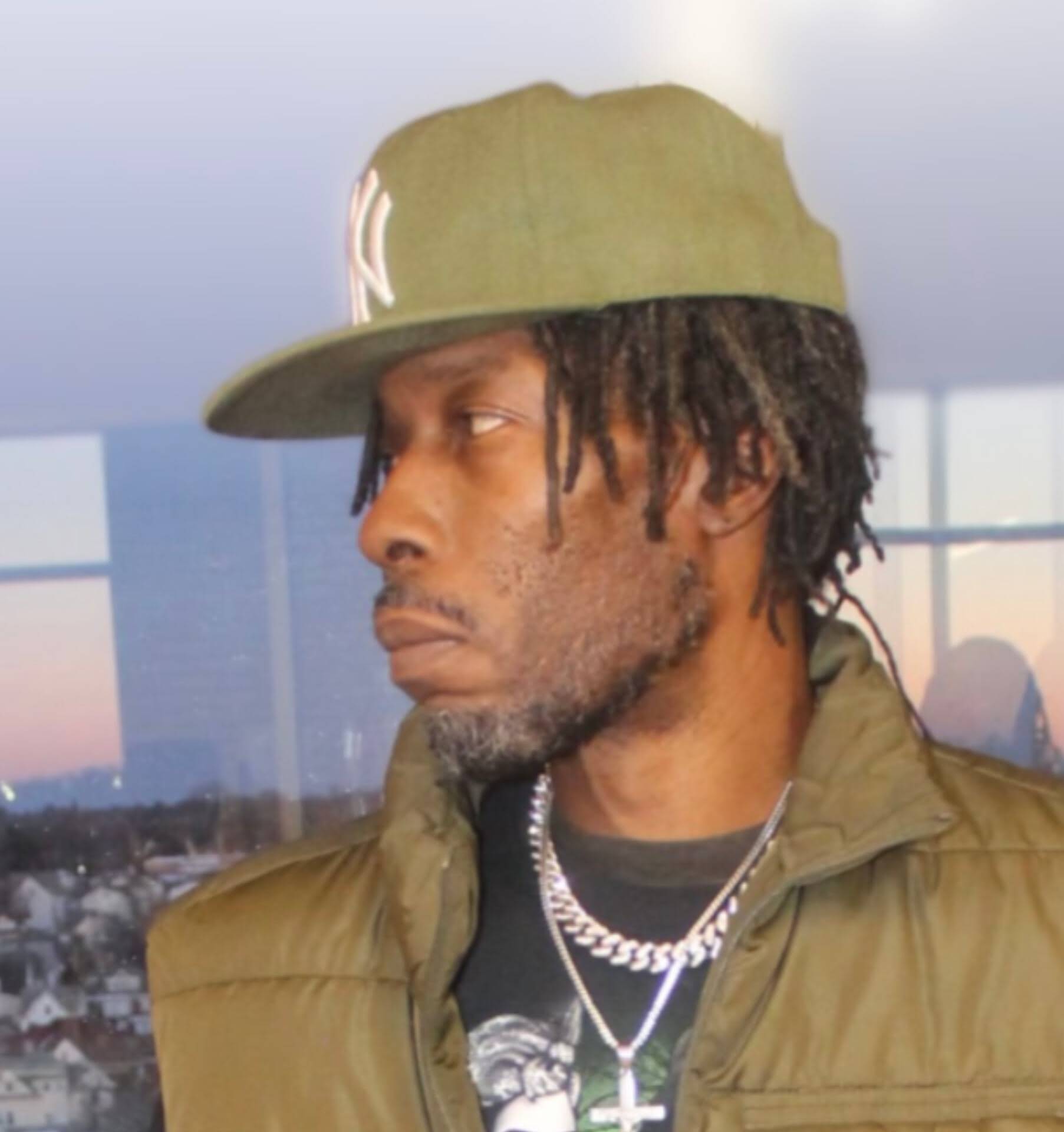 MINDBENDA in profile wearing an olive NY cap against a dusk city skyline